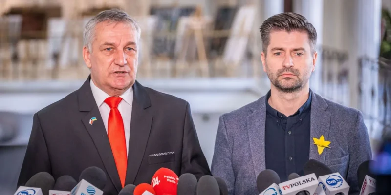 Dwóch mężczyzn w garniturach podczas konferencji prasowej stojących przed mikrofonami, po lewej z czerwonym krawatem i pinem z flagą Polski, po prawej z żółtą przypinką w kształcie gwiazdki.