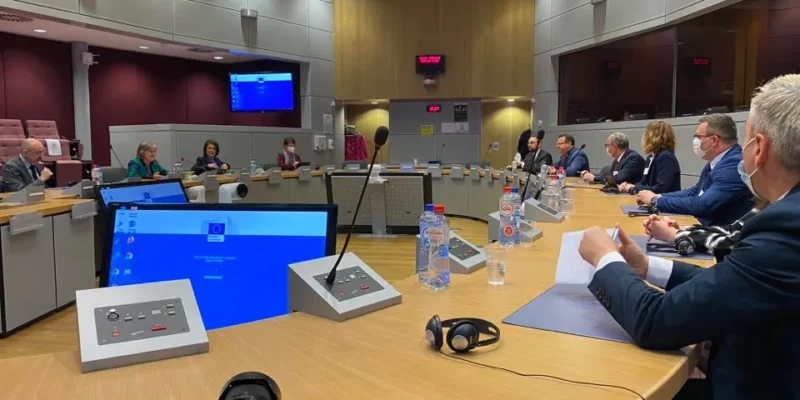 Sala konferencyjna z okrągłym stołem, grupa osób w formalnych ubraniach prowadzi dyskusję, a na pierwszym planie widać aparat, mikrofony i butelki z wodą.