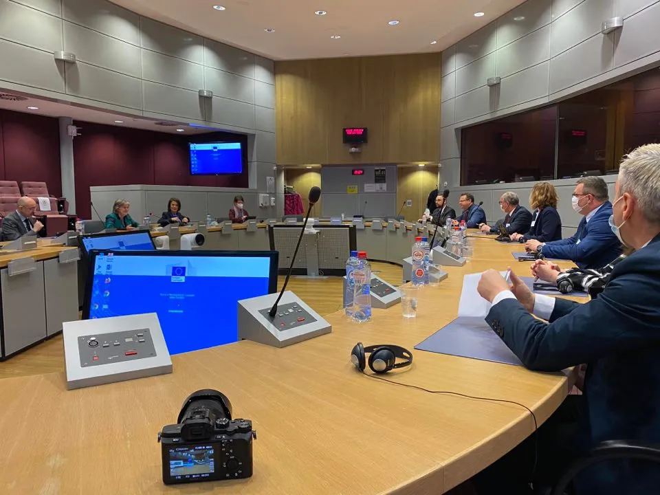 Sala konferencyjna z okrągłym stołem, grupa osób w formalnych ubraniach prowadzi dyskusję, a na pierwszym planie widać aparat, mikrofony i butelki z wodą.