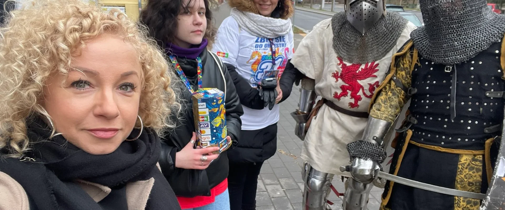 Na zdjęciu widoczna jest grupa osób pozująca do zdjęcia, w tym dwaj rycerze w zbroi oraz trzy kobiety, z których jedna trzyma opakowanie, na tle miejskiej scenerii.