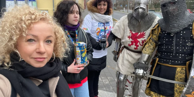 Na zdjęciu widoczna jest grupa osób pozująca do zdjęcia, w tym dwaj rycerze w zbroi oraz trzy kobiety, z których jedna trzyma opakowanie, na tle miejskiej scenerii.