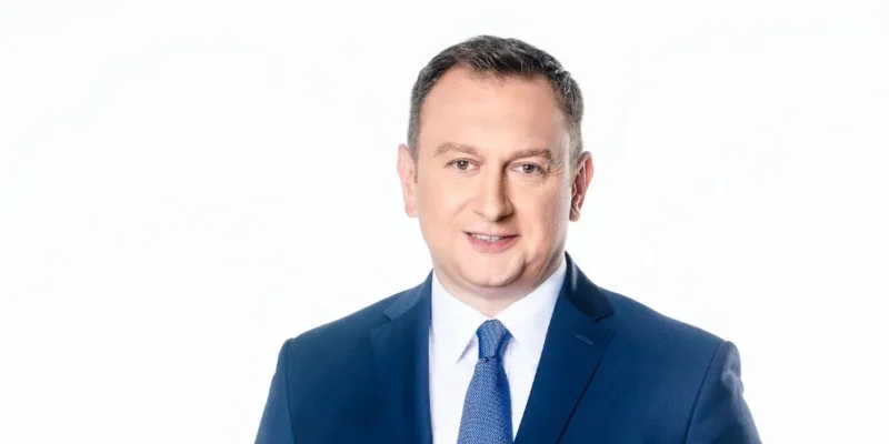 Portret mężczyzny w granatowym garniturze, białej koszuli i niebieskim krawacie na białym tle, uśmiechający się do kamery.