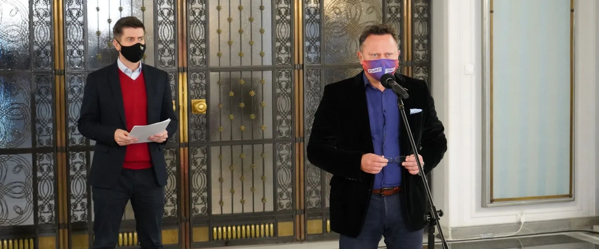 Dwóch mężczyzn w formalnych ubraniach stoi w holu z ozdobnymi metalowymi drzwiami; jeden przemawia do mikrofonu, trzymając okulary, a drugi trzyma kartki.