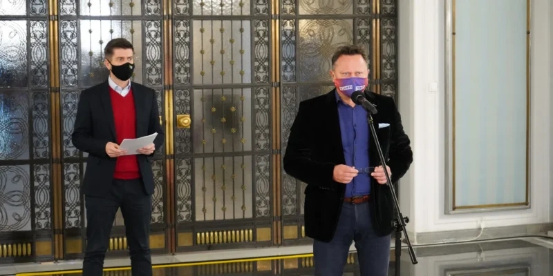 Dwóch mężczyzn w formalnych ubraniach stoi w holu z ozdobnymi metalowymi drzwiami; jeden przemawia do mikrofonu, trzymając okulary, a drugi trzyma kartki.