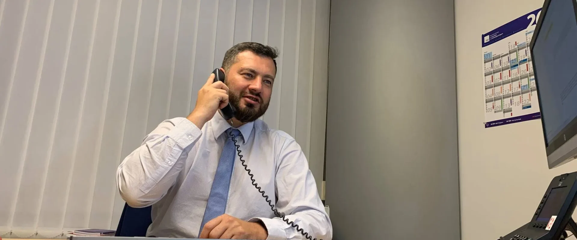 Mężczyzna w białej koszuli i niebieskim krawacie siedzi przy biurku w biurze i rozmawia przez telefon.