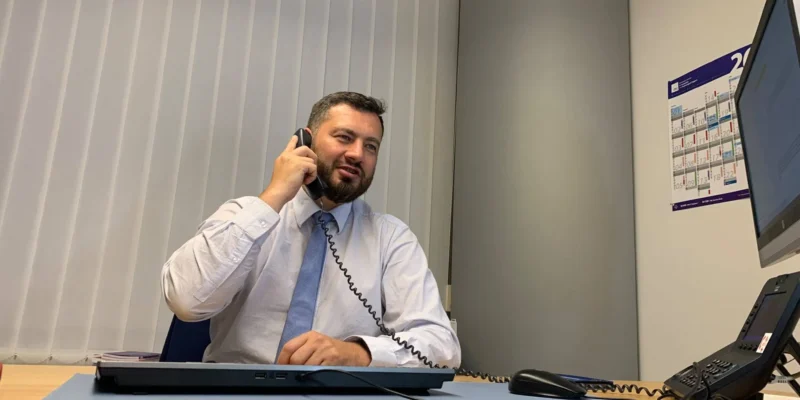 Mężczyzna w białej koszuli i niebieskim krawacie siedzi przy biurku w biurze i rozmawia przez telefon.