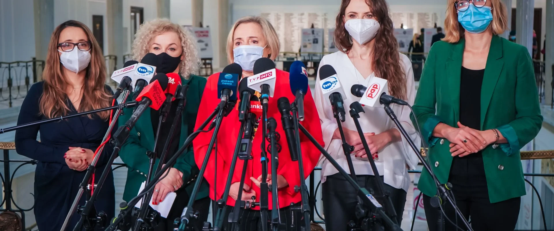 Grupa kobiet w maseczkach stoi za zestawem mikrofonów podczas konferencji prasowej w holu.