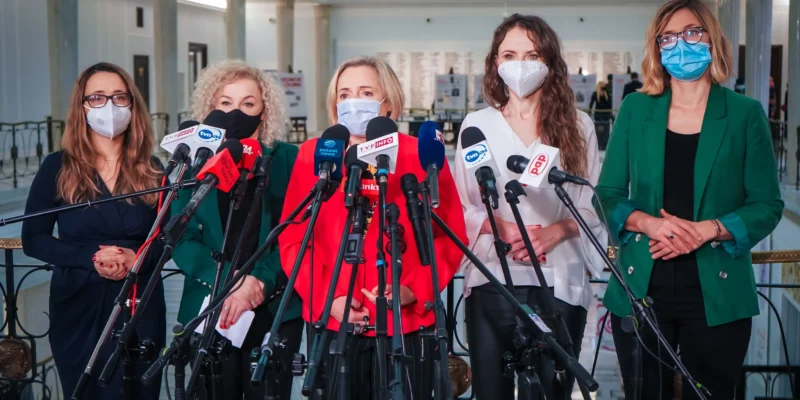 Grupa kobiet w maseczkach stoi za zestawem mikrofonów podczas konferencji prasowej w holu.