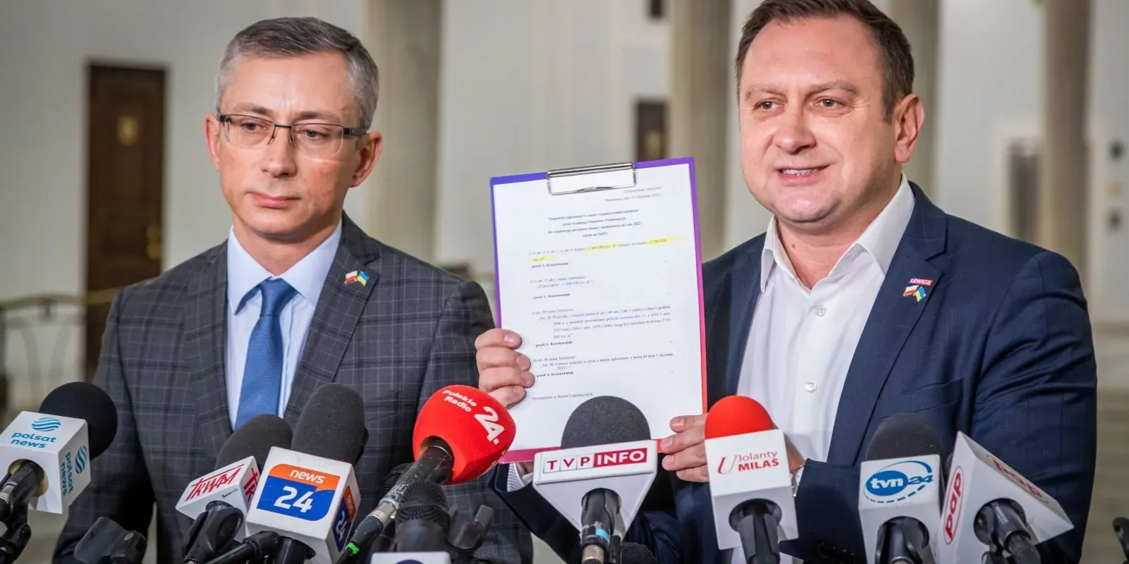 Dwóch mężczyzn w garniturach na konferencji prasowej, jeden z nich trzyma w pionie dużą kartkę z dokumentem w fioletowej oprawie, otoczeni licznymi mikrofonami mediów.