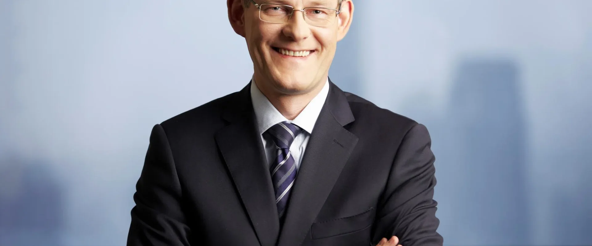 Uśmiechnięty mężczyzna w czarnym garniturze i krawacie w paski stoi z założonymi rękami na tle okna z panoramą miasta.