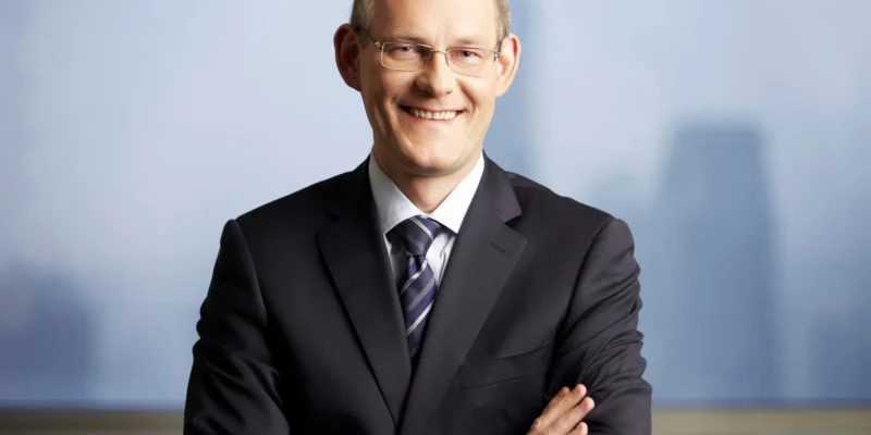 Uśmiechnięty mężczyzna w czarnym garniturze i krawacie w paski stoi z założonymi rękami na tle okna z panoramą miasta.