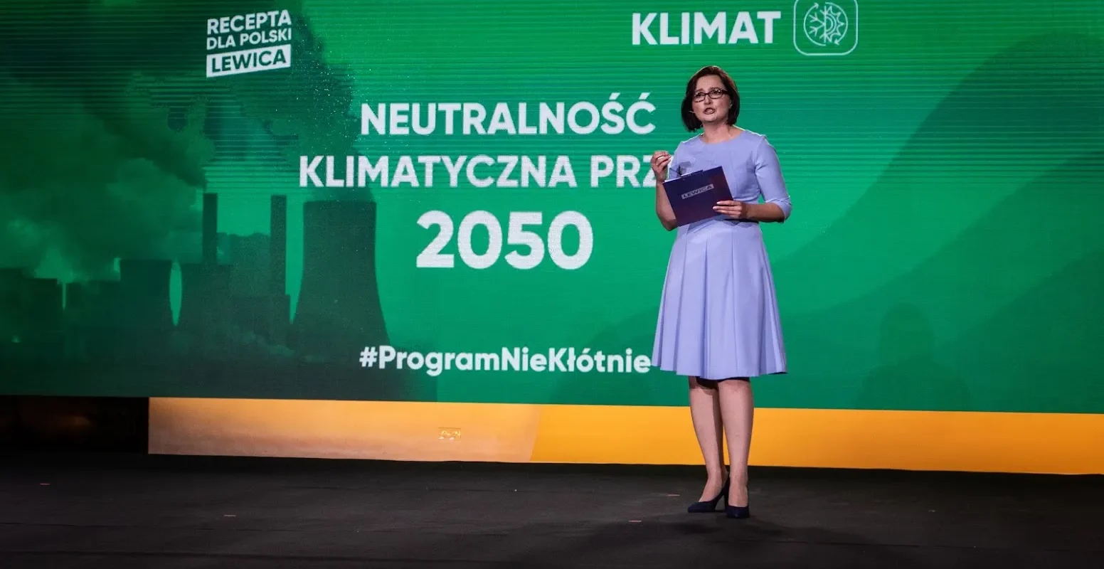 Kobieta w jasnoróżowej sukience stoi na scenie, trzyma notatnik, a w tle zielony baner z napisem o neutralności klimatycznej do 2050 i logiem Lewicy.