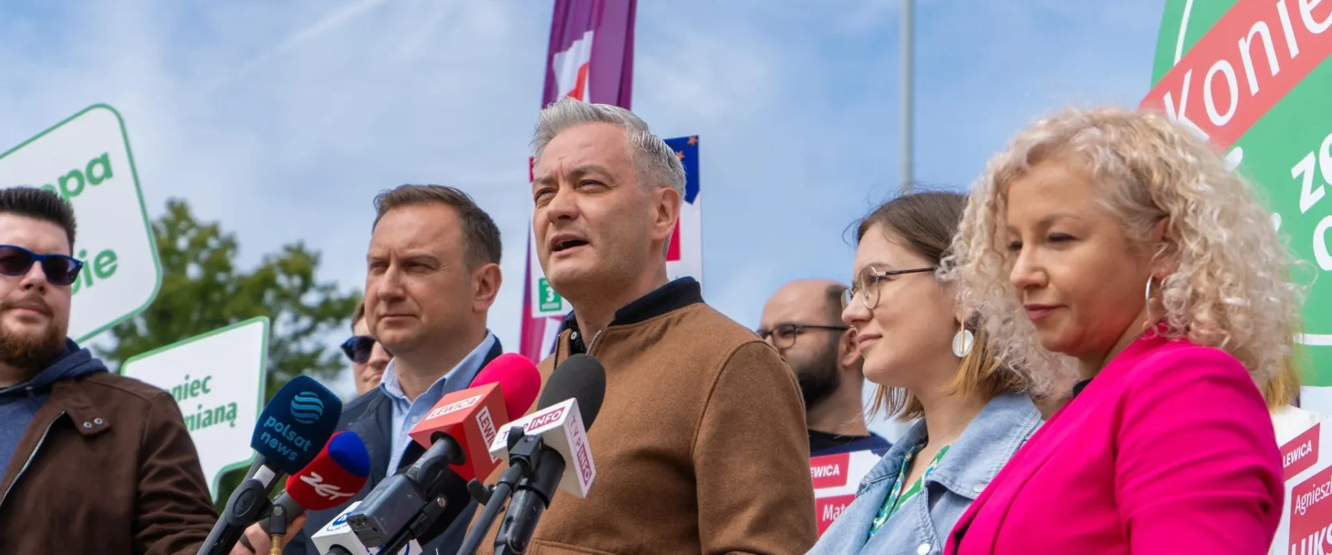 Grupa polityków podczas konferencji prasowej, centralnie stojący mężczyzna w brązowej kurtce przemawia do mediów, otoczony mikrofonami i plakatami kampanii w tle.