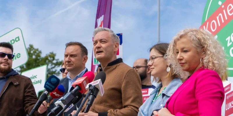 Grupa polityków podczas konferencji prasowej, centralnie stojący mężczyzna w brązowej kurtce przemawia do mediów, otoczony mikrofonami i plakatami kampanii w tle.
