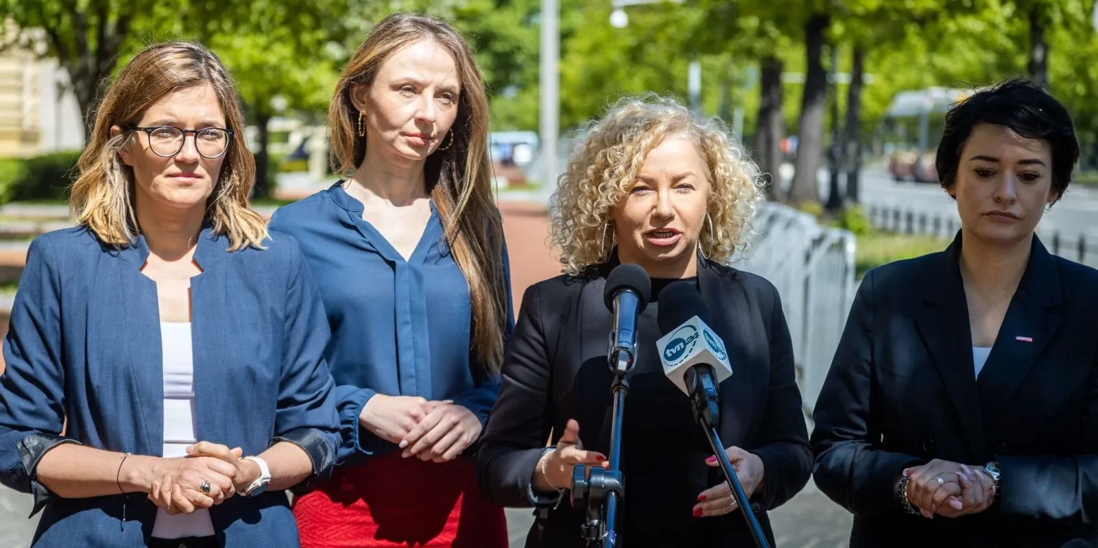 Na zdjęciu cztery kobiety stoją przed mikrofonem podczas konferencji prasowej na tle zieleni.