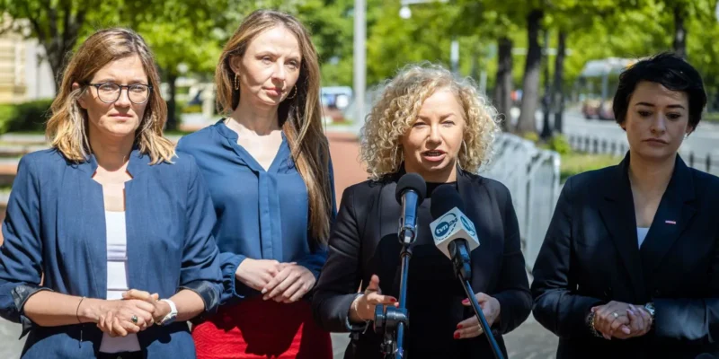 Na zdjęciu cztery kobiety stoją przed mikrofonem podczas konferencji prasowej na tle zieleni.