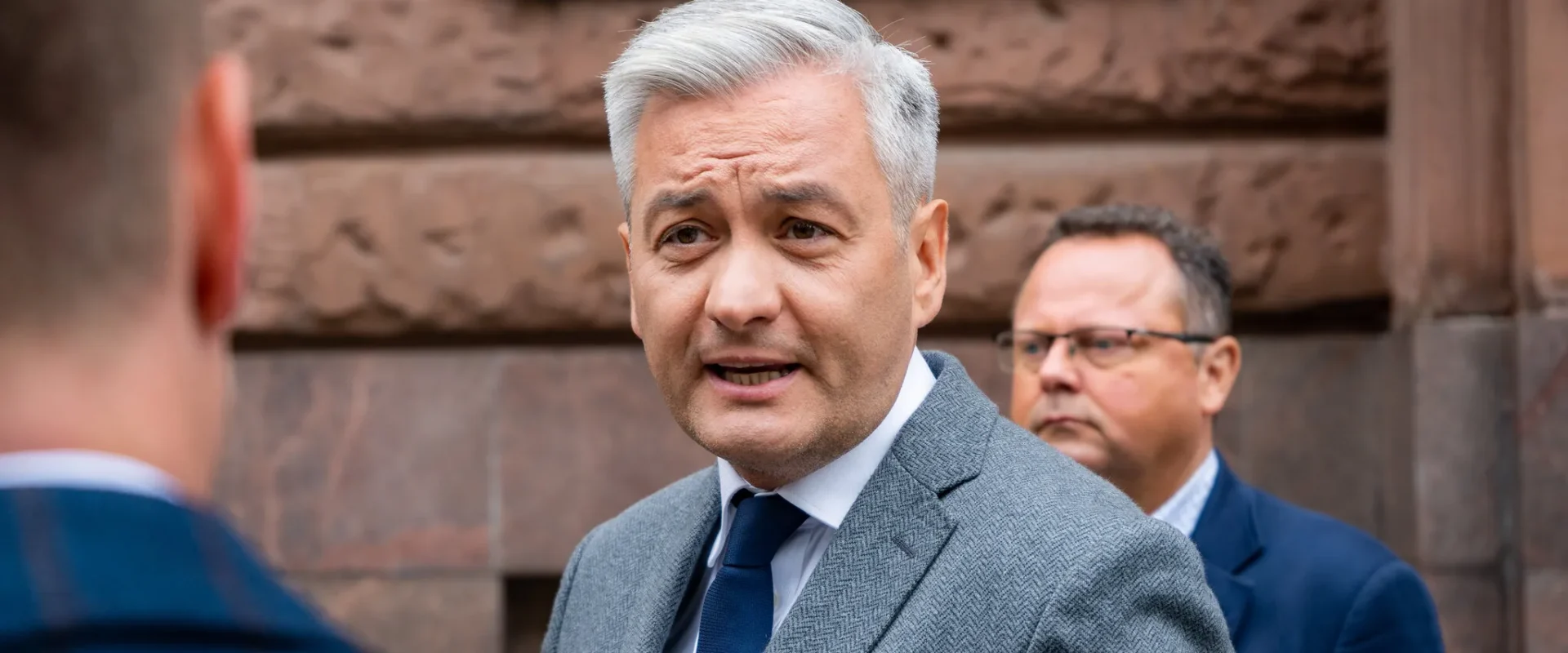 Mężczyzna w szarym garniturze rozmawia z kimś na tle historycznej, kamiennej ściany.