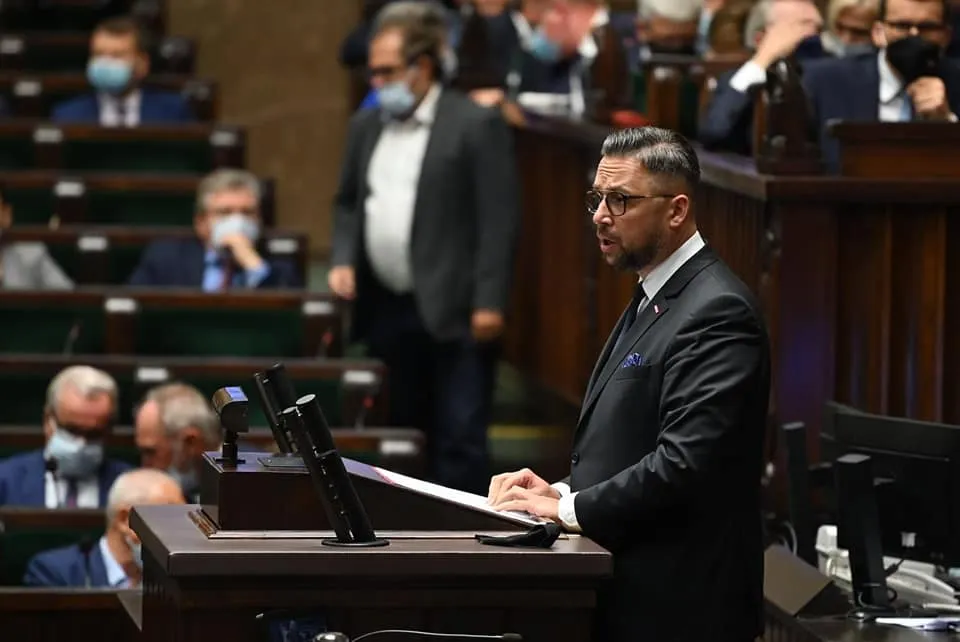 Mężczyzna w garniturze przemawia z mównicy w sali plenarnej parlamentu, a w tle widoczni są posłowie, część z nich w maseczkach.