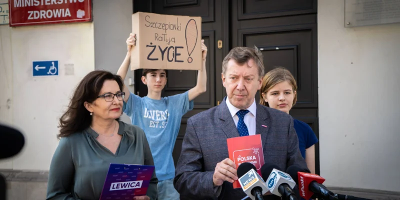 Mężczyzna w garniturze przemawia do mikrofonów przed wejściem do budynku Ministerstwa Zdrowia; za nim osoba trzyma karton z napisem „Szczepionki ratują ŻYCIE!”, a obok stoi kobieta z teczką Lewica.