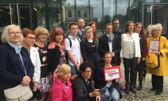 Kilkunastoosobowa grupa stoi przed szklaną elewacją budynku, część osób trzyma tabliczki z napisem „Ratujmy Kobiety” po złożeniu podpisów pod rejestracją komitetu Ratujmy Kobiety 2017.