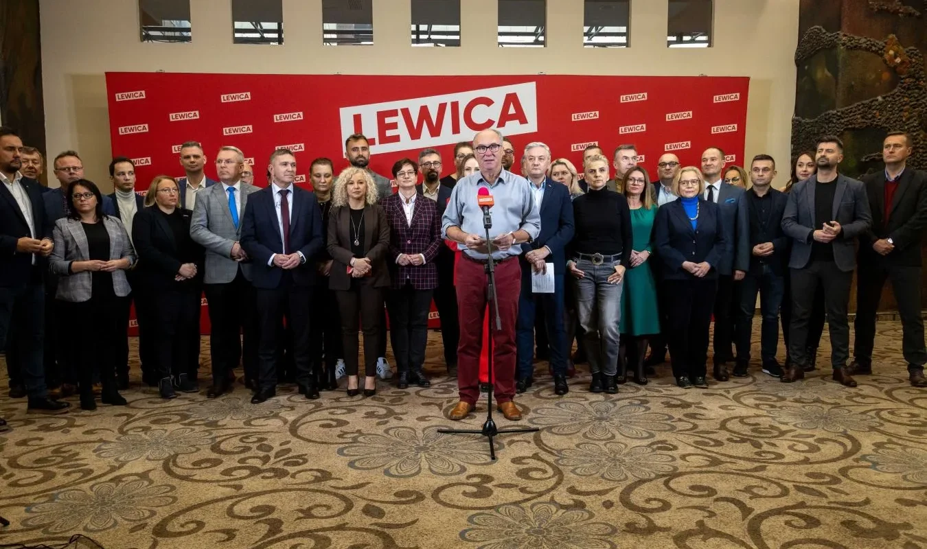 Grupa polityków Lewicy stoi na konferencji prasowej w sali, przed czerwonym tłem z napisem LEWICA, a mężczyzna przy mikrofonie wygłasza oświadczenie o porozumieniu powyborczym mającym doprowadzić do utworzenia wspólnego rządu.