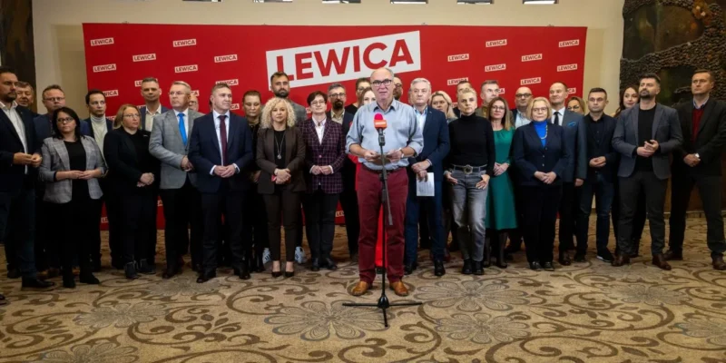 Grupa polityków Lewicy stoi na konferencji prasowej w sali, przed czerwonym tłem z napisem LEWICA, a mężczyzna przy mikrofonie wygłasza oświadczenie o porozumieniu powyborczym mającym doprowadzić do utworzenia wspólnego rządu.