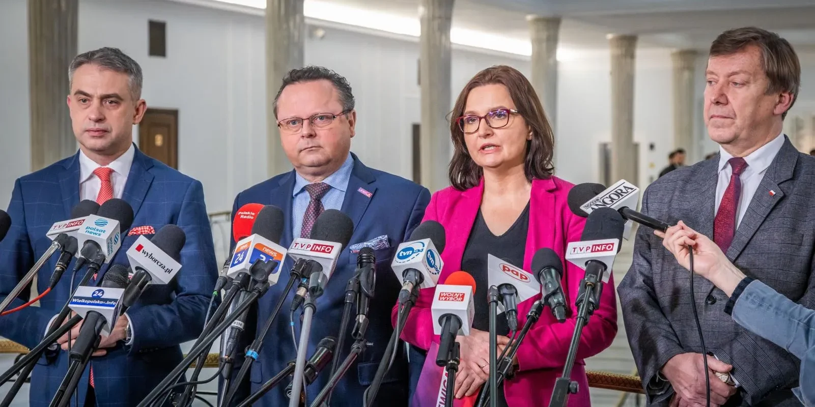 Na zdjęciu cztery osoby w garniturach stoją przed mikrofonami, wypowiadając się podczas konferencji prasowej.