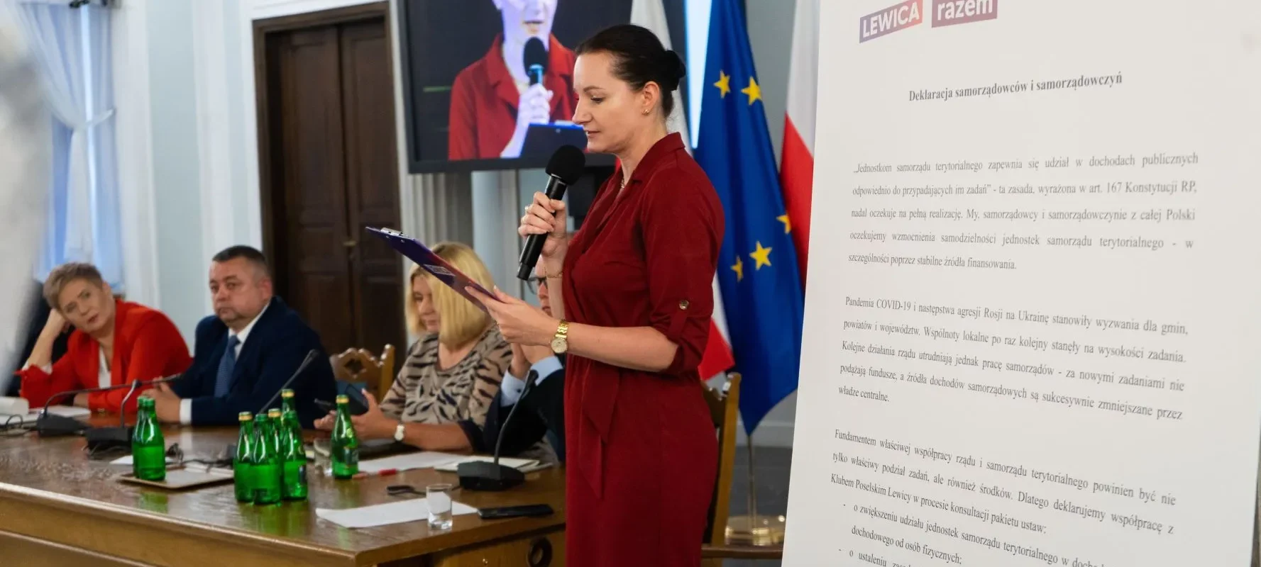 Na zdjęciu widoczna jest kobieta w czerwonej sukience, która przemawia do zebranych na konferencji, obok niej siedzą inni uczestnicy spotkania, w tle widoczne są flagi oraz dokumenty dotyczące samorządności.