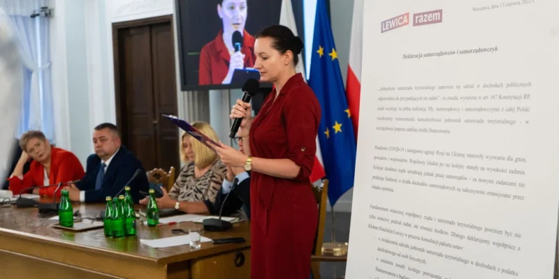 Na zdjęciu widoczna jest kobieta w czerwonej sukience, która przemawia do zebranych na konferencji, obok niej siedzą inni uczestnicy spotkania, w tle widoczne są flagi oraz dokumenty dotyczące samorządności.