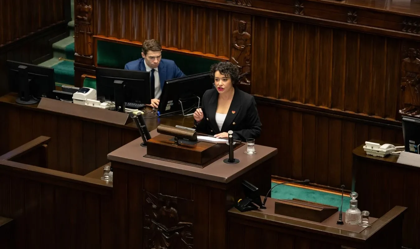 Kobieta w czarnym garniturze przemawia z mównicy w sali plenarniej, podczas gdy mężczyzna siedzi za nią przy dwóch monitorach.