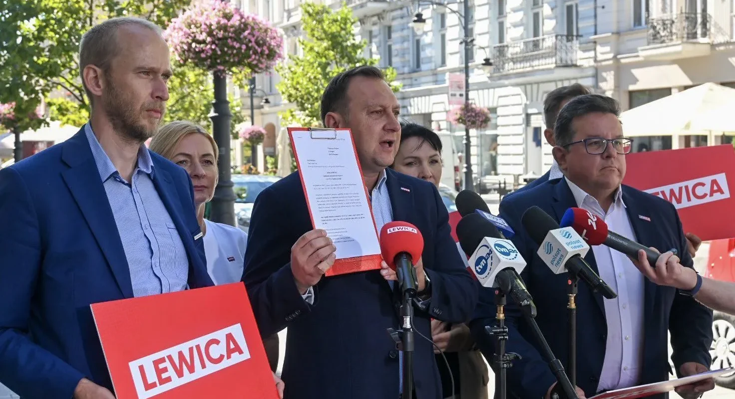 Grupa osób podczas konferencji prasowej na ulicy; mężczyzna w garniturze trzyma dokumenty w czerwonej teczce i stoi przy mikrofonach, a w tle widać plakaty Lewicy.