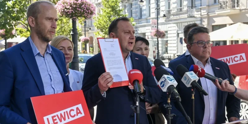 Grupa osób podczas konferencji prasowej na ulicy; mężczyzna w garniturze trzyma dokumenty w czerwonej teczce i stoi przy mikrofonach, a w tle widać plakaty Lewicy.