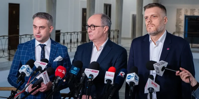 Trzech mężczyzn w garniturach stoi przed mikrofonami i udziela informacji w przestrzeni publicznej.