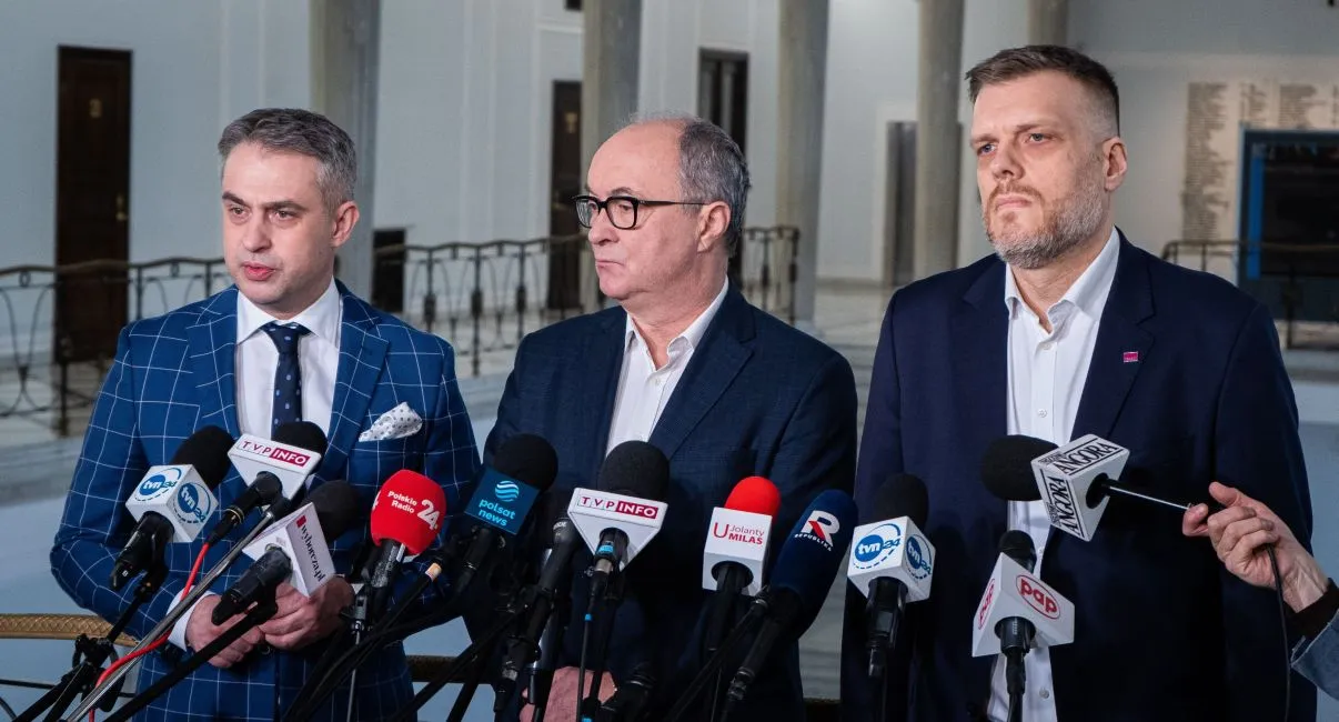 Trzech mężczyzn w garniturach stoi przed mikrofonami i udziela informacji w przestrzeni publicznej.