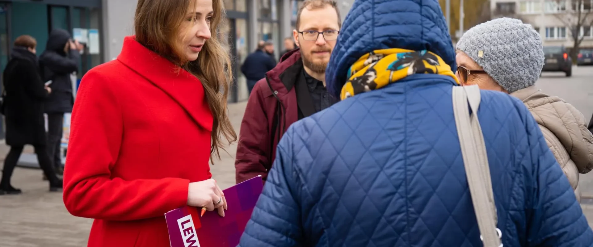 Kobieta w czerwonym płaszczu trzyma plakat z napisem Lewica i rozmawia z innymi osobami na miejskim chodniku.