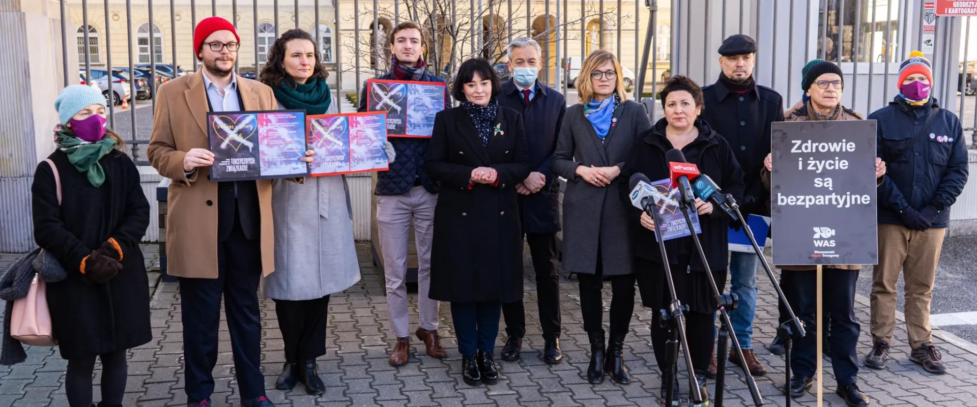 Grupa osób w zimowych ubraniach stoi na zewnątrz przed ogrodzeniem budynku, trzyma plakaty i mikrofony podczas akcji promującej 7 pomysłów na lepsze powietrze w Warszawie.
