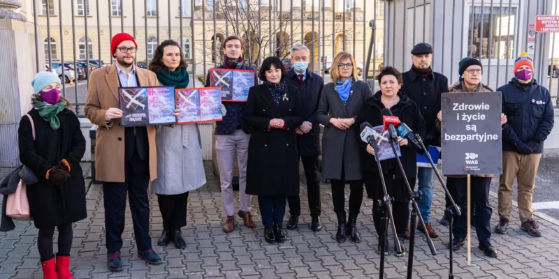 Grupa osób w zimowych ubraniach stoi na zewnątrz przed ogrodzeniem budynku, trzyma plakaty i mikrofony podczas akcji promującej 7 pomysłów na lepsze powietrze w Warszawie.
