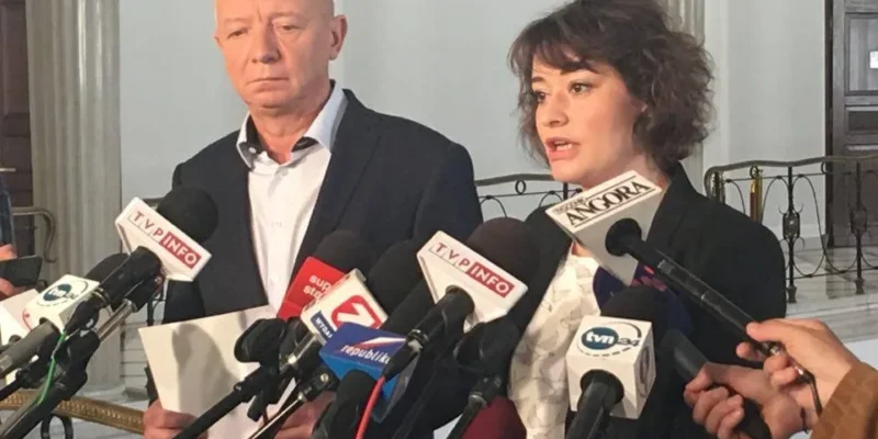 Dwie osoby przemawiają do mediów przy stojaku z licznymi mikrofonami w korytarzu Sejmu.