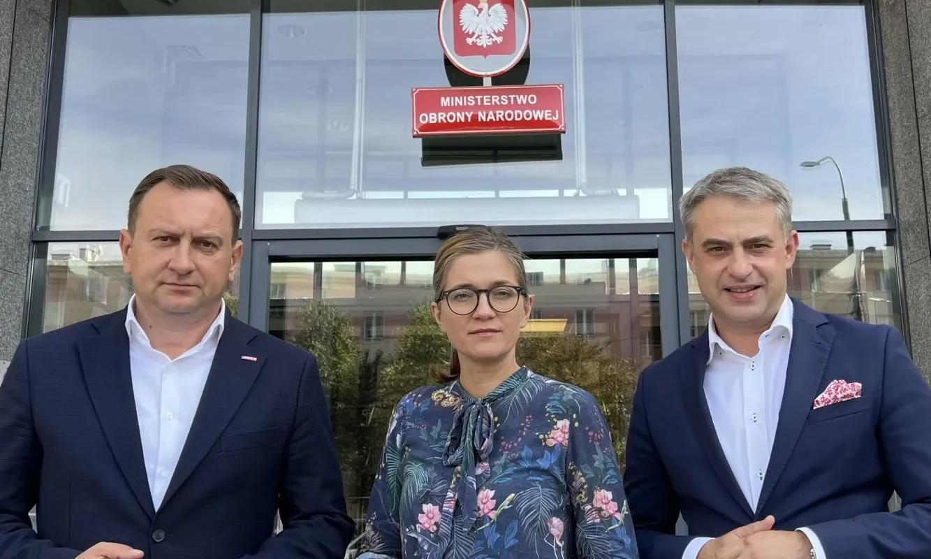 Trzy osoby stoją przed wejściem do Ministerstwa Obrony Narodowej; w centrum kobieta w okularach i kwiecistej bluzce, po bokach mężczyźni w garniturach, nad wejściem widnieje godło Polski i tabliczka z napisem Ministerstwo Obrony Narodowej.