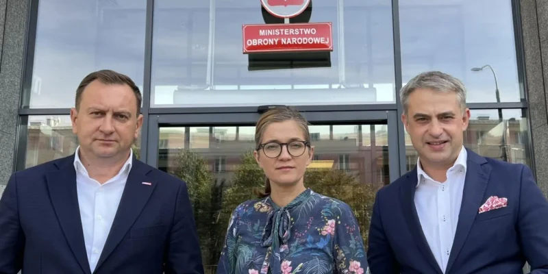 Trzy osoby stoją przed wejściem do Ministerstwa Obrony Narodowej; w centrum kobieta w okularach i kwiecistej bluzce, po bokach mężczyźni w garniturach, nad wejściem widnieje godło Polski i tabliczka z napisem Ministerstwo Obrony Narodowej.