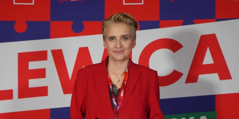 Kobieta w czerwonym garniturze stoi na kolorowym tle w puzzle’owym wzorze z dużym napisem LEWICA, na szyi ma identyfikator.