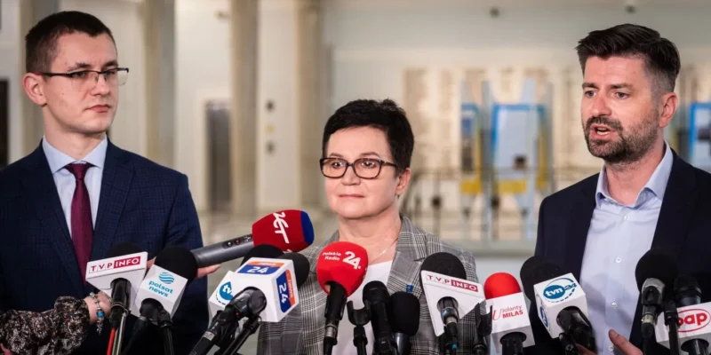 Trzy osoby stoją na konferencji prasowej za licznymi mikrofonami różnych stacji telewizyjnych, z kobietą w okularach w centrum i dwoma mężczyznami po bokach.