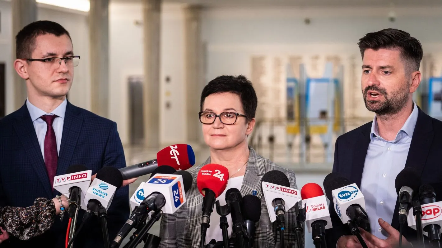 Trzy osoby stoją na konferencji prasowej za licznymi mikrofonami różnych stacji telewizyjnych, z kobietą w okularach w centrum i dwoma mężczyznami po bokach.