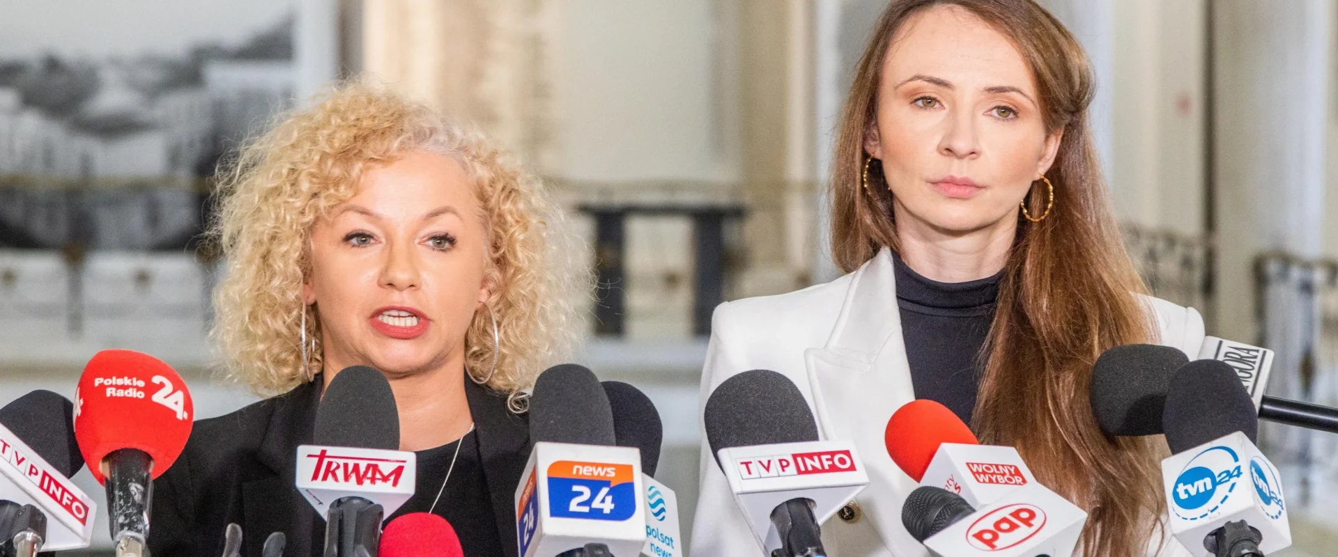 Dwie kobiety stoją przed licznymi mikrofonami dziennikarzy podczas konferencji prasowej w korytarzu, blond kobieta po lewej mówi, a brunetka po prawej nosi białą marynarkę i obserwuje.