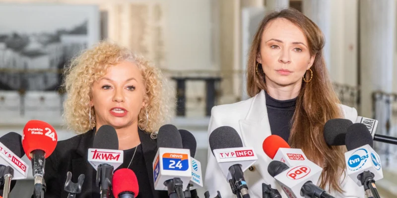 Dwie kobiety stoją przed licznymi mikrofonami dziennikarzy podczas konferencji prasowej w korytarzu, blond kobieta po lewej mówi, a brunetka po prawej nosi białą marynarkę i obserwuje.