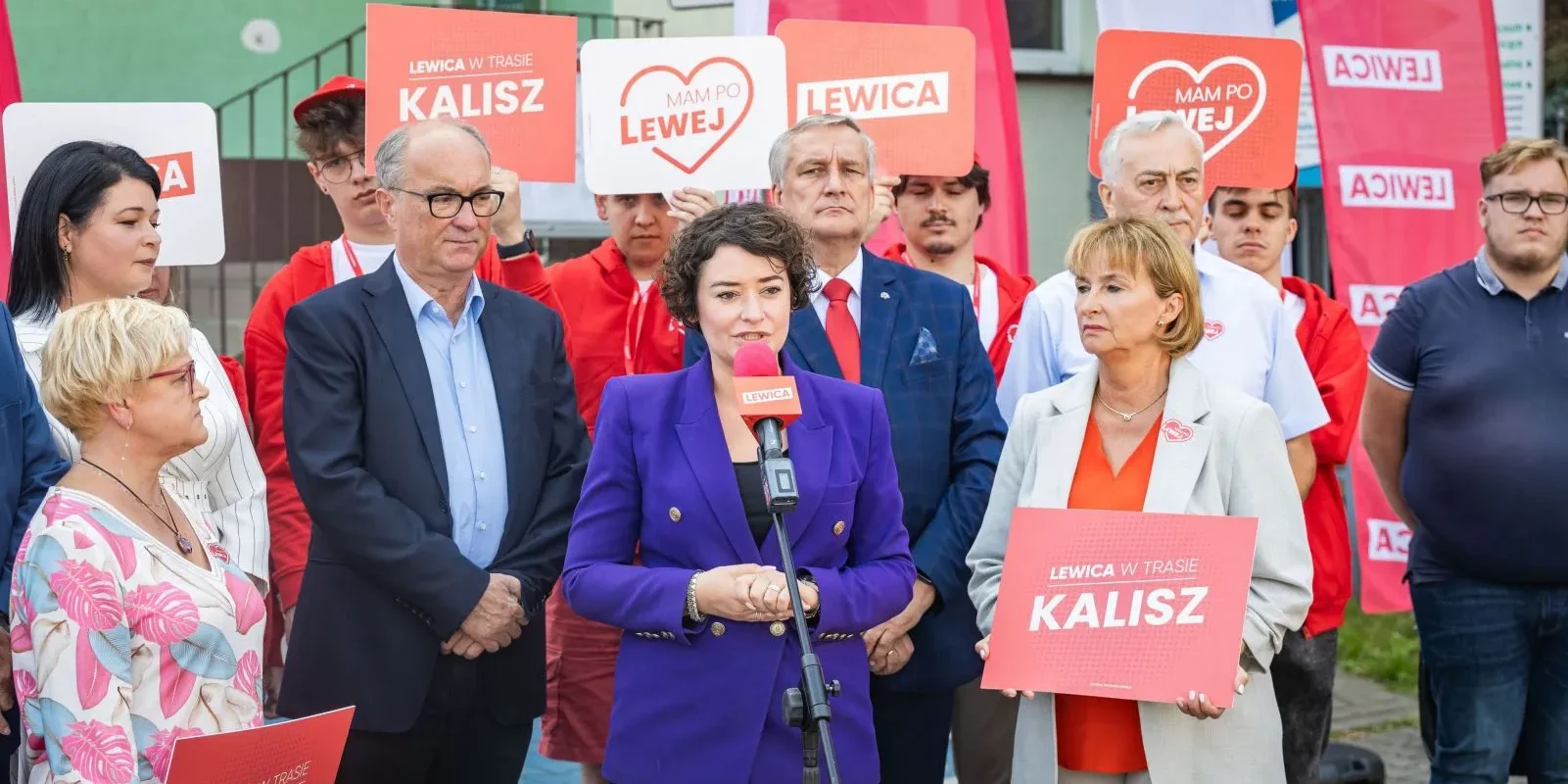 Grupa polityków Lewicy podczas konferencji prasowej na ulicy, w centrum kobieta w fioletowej marynarce przemawia do mikrofonu, w tle plakaty z napisem Lewica i Kalisz.
