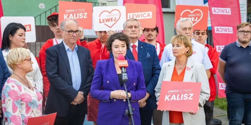Grupa polityków Lewicy podczas konferencji prasowej na ulicy, w centrum kobieta w fioletowej marynarce przemawia do mikrofonu, w tle plakaty z napisem Lewica i Kalisz.