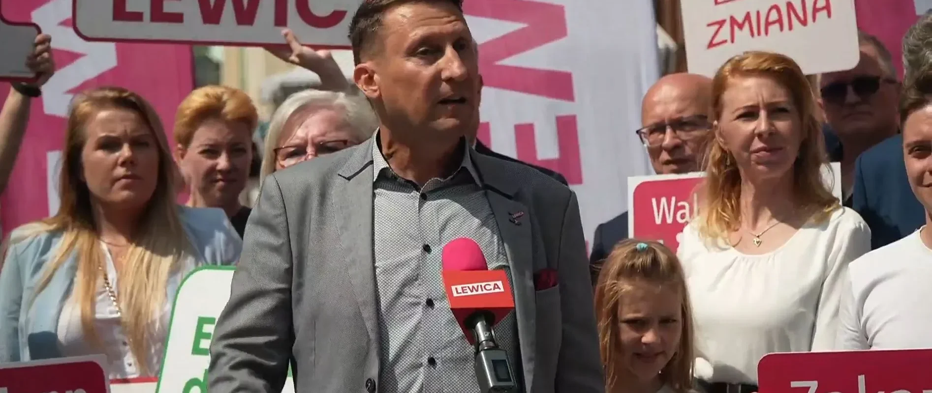 Mężczyzna w garniturze przemawia do tłumu na konferencji, trzymając mikrofon z napisem LEWICA; w tle stoją zwolennicy z plakatami i banerami Lewicy.