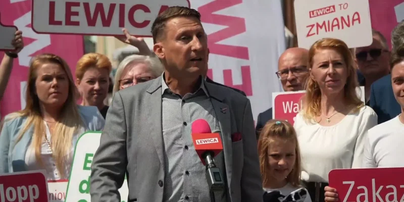 Mężczyzna w garniturze przemawia do tłumu na konferencji, trzymając mikrofon z napisem LEWICA; w tle stoją zwolennicy z plakatami i banerami Lewicy.