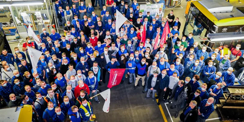 Grupa pracowników w fabryce, ubranych w niebieskie koszulki i maski, z flagami i transparentami, gromadzi się w hali produkcyjnej.